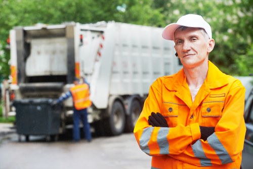A professional waste management worker in a high-visibility orange jacket with reflective grey and yellow stripes is standing outdoors with arms crossed, smiling slightly. Behind him, a large white rubbish collection vehicle is parked on a driveway, with its rear compartment open, revealing an empty compartment ready for waste loading. The scene is set in a leafy area with green trees and bushes in the background, typical of a residential or semi-urban setting. To the left, another worker in a similar uniform is seen near a black wheeled bin, possibly preparing for rubbish collection or clearance. The environment appears clean, well-maintained, and well-lit with natural daylight, emphasizing the professional and organized approach to rubbish removal services provided by House Clearance Hertford, covering local waste disposal needs in the Hertford area. The overall scene conveys a clear focus on efficient rubbish collection and clearance operations, aligning with local waste management services in the Hertford postcode area.