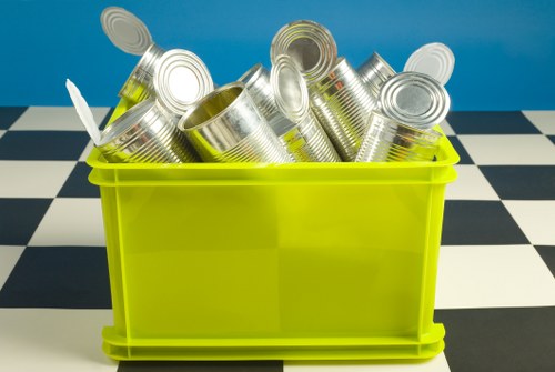 The image shows a bright yellow plastic bin filled with empty, silver tin cans of varying sizes, some with visible ridges and pull-tabs, placed on a black and white checkered floor. The background is a solid blue wall, and the cans are stacked in a somewhat disorganized manner, with some leaning against others and slightly spilling over the top edge of the bin. This scene appears to depict a collection of recyclable metal waste, typical of rubbish removal or house clearance activities in Hertford. The overall setting suggests an indoor or outdoor space designated for waste sorting or collection, with the focus on the metallic cans that are ready for removal or recycling by a service such as House Clearance Hertford.