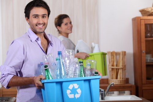 A young man with dark hair and a beard, smiling while holding a blue recycling bin filled with empty glass bottles and jars, set in a kitchen environment. The bin features a white recycling symbol on the side and is partially visible, with the man standing near a kitchen sink and countertop. In the background, a woman with dark hair pulled back is seen standing upright, looking to the side, with a green storage box and a wooden cabinet behind her, along with a basket and some paper towels. The kitchen has light-colored walls and wooden cupboards, creating a tidy and organized setting. The scene suggests a household waste collection or rubbish removal activity, consistent with services offered by House Clearance Hertford, located in Hertford. The lighting appears natural, emphasizing the textures of the glass bottles and the kitchen furnishings, supporting the context of domestic rubbish disposal or recycling efforts.