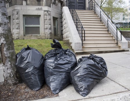 Three black plastic rubbish bags, filled and tied at the top, are placed on the pavement beside a tree in front of a building with a stone staircase leading up to an entrance with metal railings. The bags appear to contain household waste and are positioned close to the curb, likely for collection. The background includes a stone building with small windows and a grassy area at the base of the tree. The scene is outdoors, with natural daylight illuminating the area, typical of rubbish disposal and removal steps in Hertford. This image exemplifies waste ready for rubbish collection services, such as those offered by House Clearance Hertford, serving local residential properties and ensuring tidy outdoor spaces.