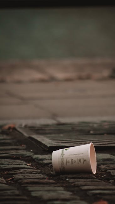A collection of overflowing rubbish bins and scattered waste bags on a paved urban street corner, with various black, red, and green bins used for mixed refuse, paper, and recyclable items. The waste 