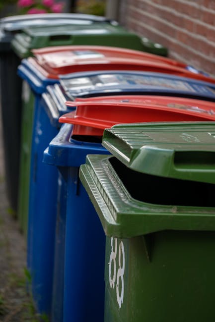 A row of large, rectangular wheelie bins positioned along a paved pathway beside a red brick wall. The bins are painted in various colors, including green, red, and blue, with textured lids featuring
